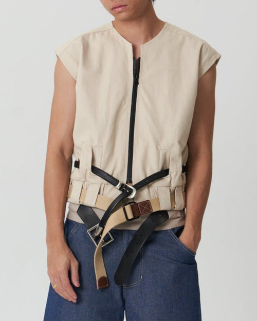 A person wearing a beige sleeveless vest with a black YKK zipper closure and belt loops at the waist. They are also wearing blue jeans.